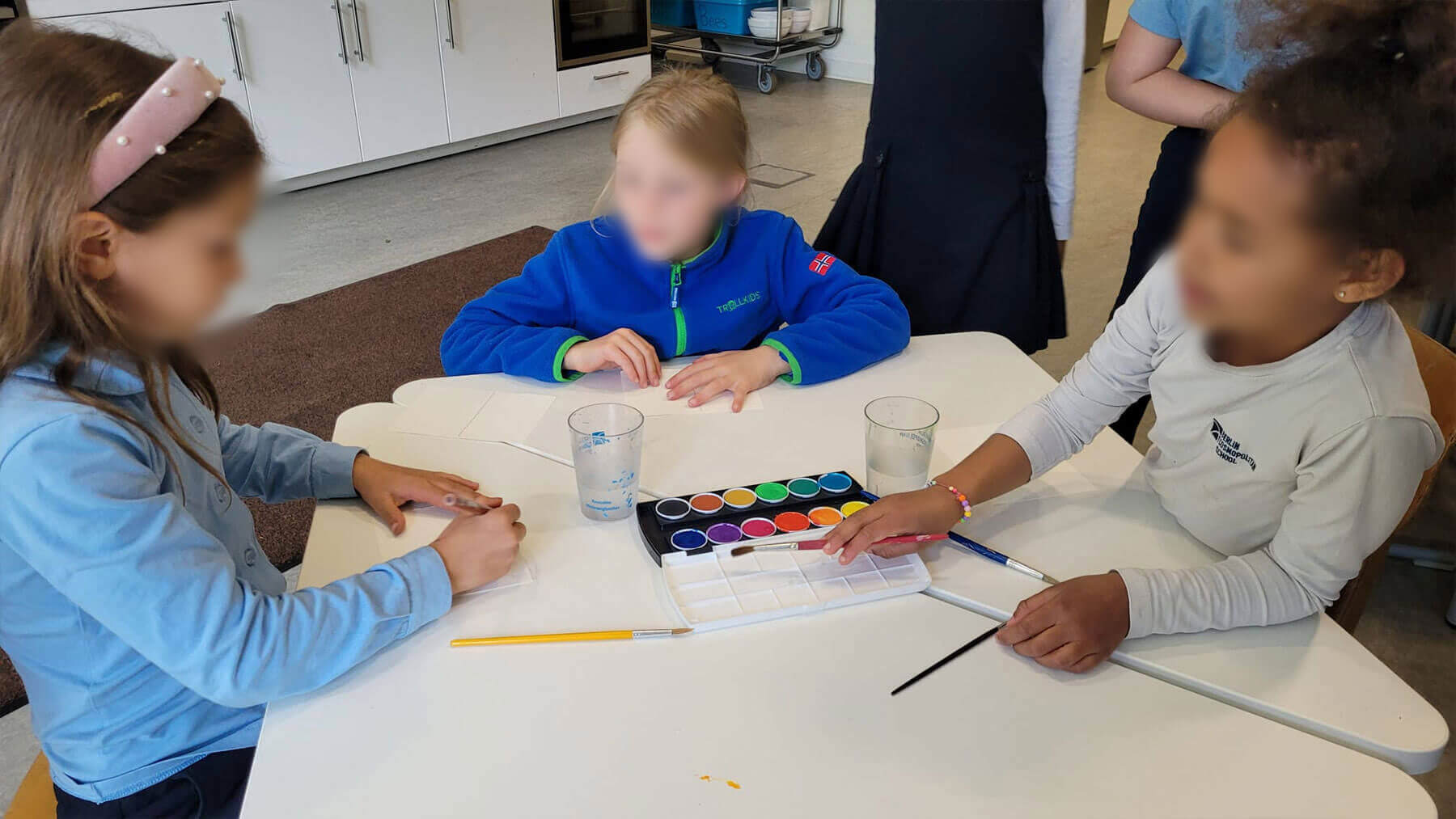 Cosmopolitanschool Berlin Drei Kinder sitzen um einen weißen Tisch mit Aquarellfarben, Pinseln und Papier und bereiten sich in einem hell erleuchteten Klassenzimmer auf das gemeinsame Malen vor. Ihre Gesichter sind aus Gründen der Privatsphäre unscharf.