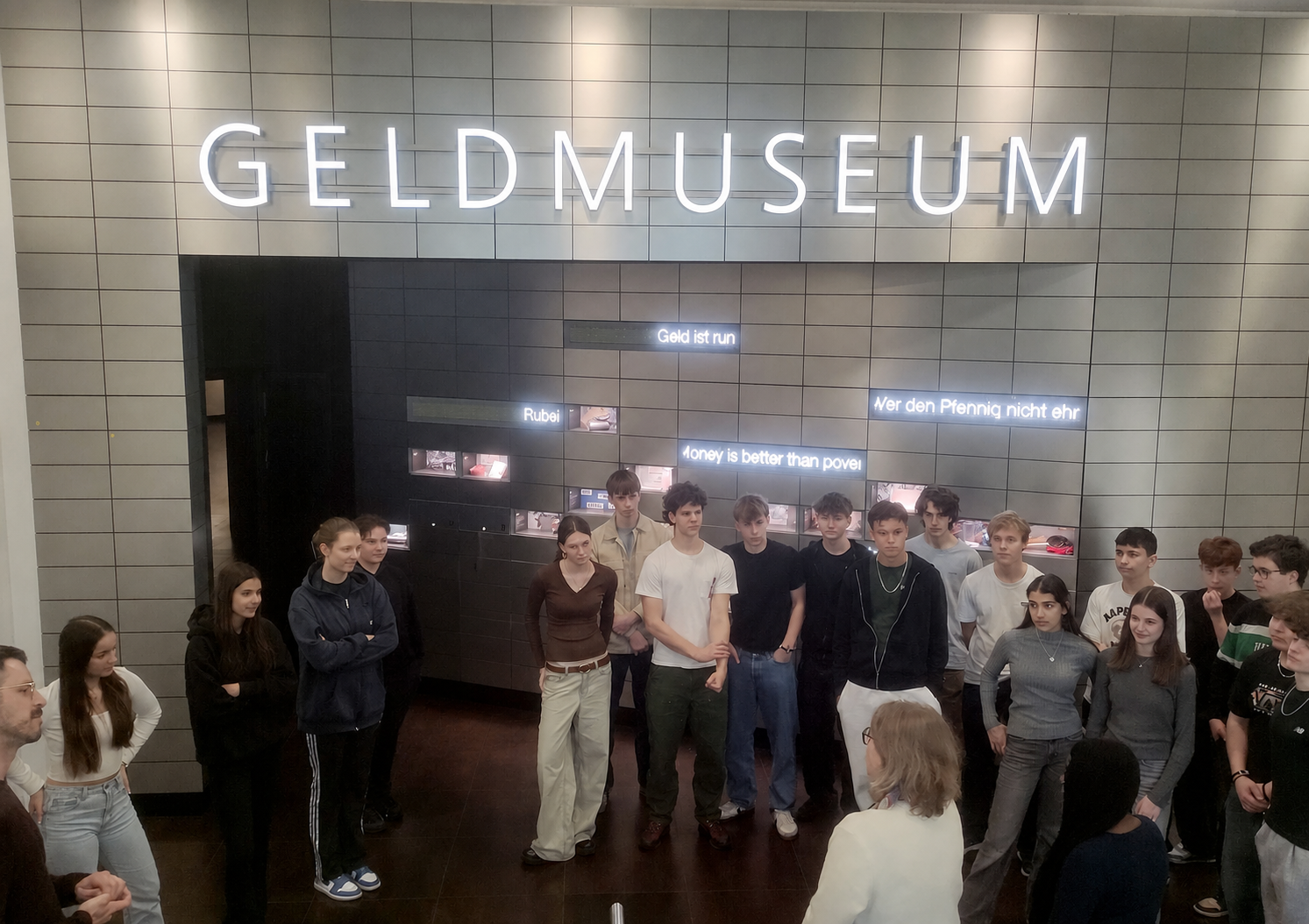 Gruppe junger Menschen steht vor einer Wand mit der Aufschrift 'GELDMUSEUM' in großen Leuchtbuchstaben. mit KI bearbeitet