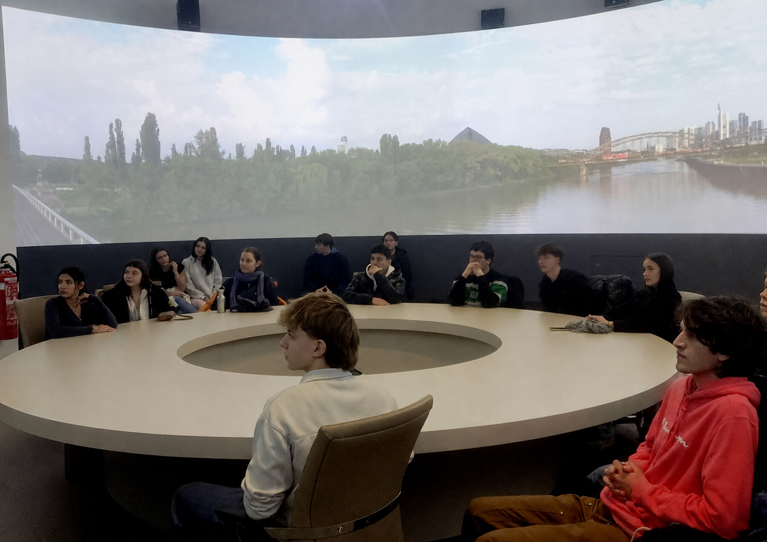 Jugendliche sitzen an einem runden Tisch vor einer großen Panorama-Leinwand mit Fluss- und Stadtlandschaft. mit KI bearbeitet