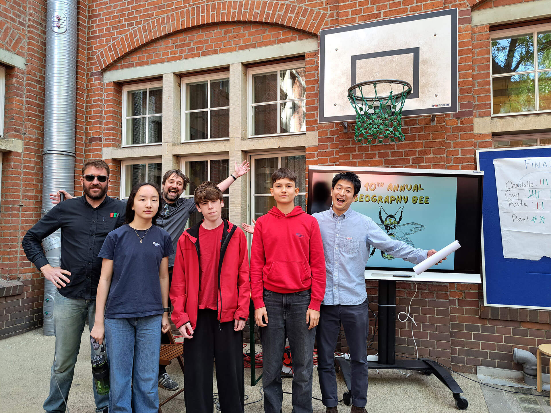 Cosmopolitanschool Berlin Sechs Personen posieren vor einer Backsteinmauer mit Fenstern, einem Basketballkorb und einem Bildschirm mit der Aufschrift „Geography Bee“. Drei Schüler stehen davor, zwei Erwachsene dahinter und eine Person posiert energisch seitlich.