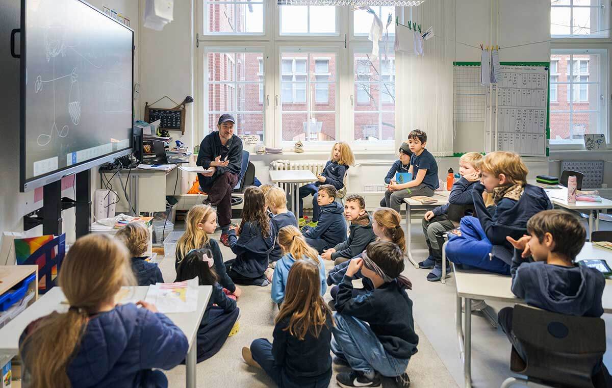 Cosmopolitanschool Berlin Ein Lehrer sitzt vorne in einem Klassenzimmer und spricht zu einer Gruppe aufmerksamer Grundschüler, die auf dem Boden und an den Tischen sitzen. Das Klassenzimmer hat große Fenster, ein digitales Whiteboard und Lernplakate. loading=