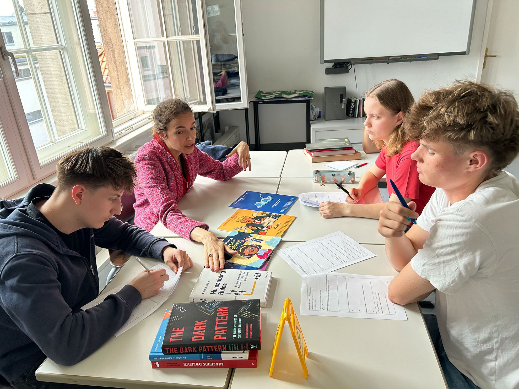 Cosmopolitanschool Berlin Eine Frau in einer rosafarbenen Jacke spricht mit drei Schülern, die in einem hellen Klassenzimmer mit großen Fenstern um einen Tisch mit Papieren und Büchern, darunter "The Dark Pattern", sitzen, während sie zuhören und sich Notizen machen. Mit KI bearbeitet.