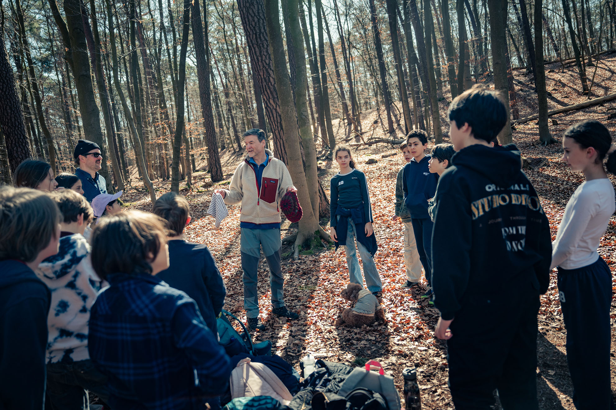 Cosmopolitanschool Berlin Eine Gruppe von Kindern steht im Kreis um einen Erwachsenen in einem Wald an der Waldschule BCS und hört aufmerksam zu. Der Boden ist mit abgefallenen Blättern bedeckt und die hohen, blattlosen Bäume lassen auf einen frühen Frühling oder einen späten Herbst schließen. loading=