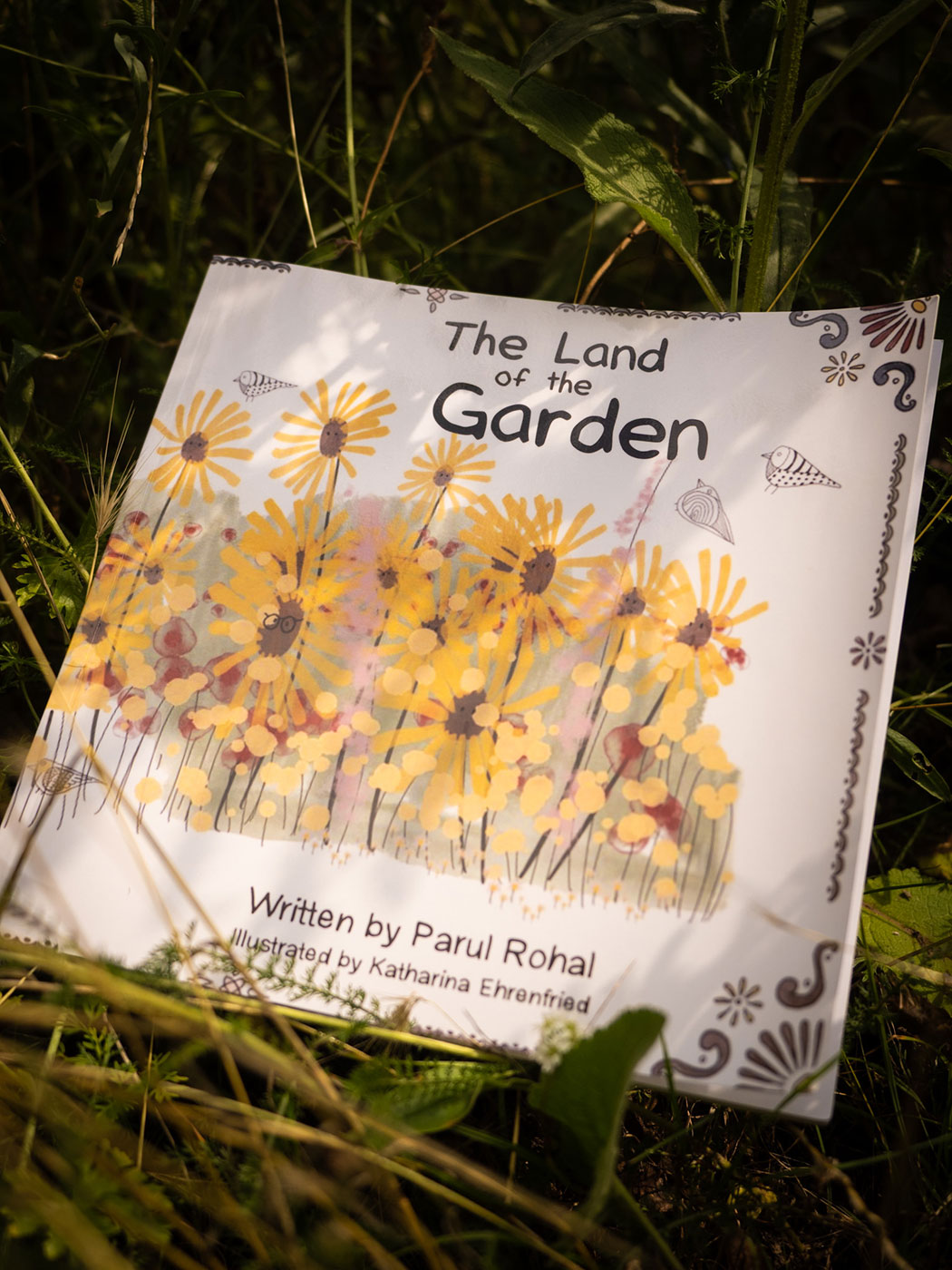 Cosmopolitanschool Berlin Ein Kinderbuch mit dem Titel Das Land des Gartens von Parul Rohal, illustriert von Katharina Ehrenfreund, liegt im Gras. Auf dem Einband sind gelbe Blumen, Bienen, einfache Zierleisten und der Hinweis "Automatisch gespeicherter Entwurf" in der Ecke zu sehen. loading=