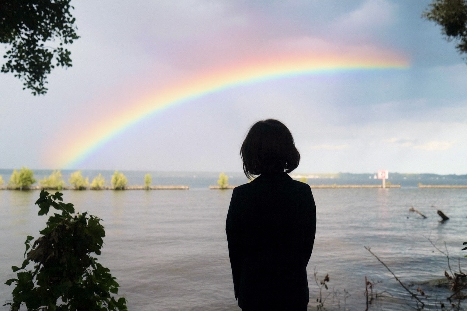 Cosmopolitanschool Berlin Eine Person mit kurzen Haaren steht am Müggelsee und blickt auf einen leuchtenden Regenbogen, der sich über einen wolkenverhangenen Himmel wölbt. Bäume rahmen das Wasser ein und schaffen eine friedliche, kontemplative Szene - die perfekte Inspiration für ein Naturseminar.