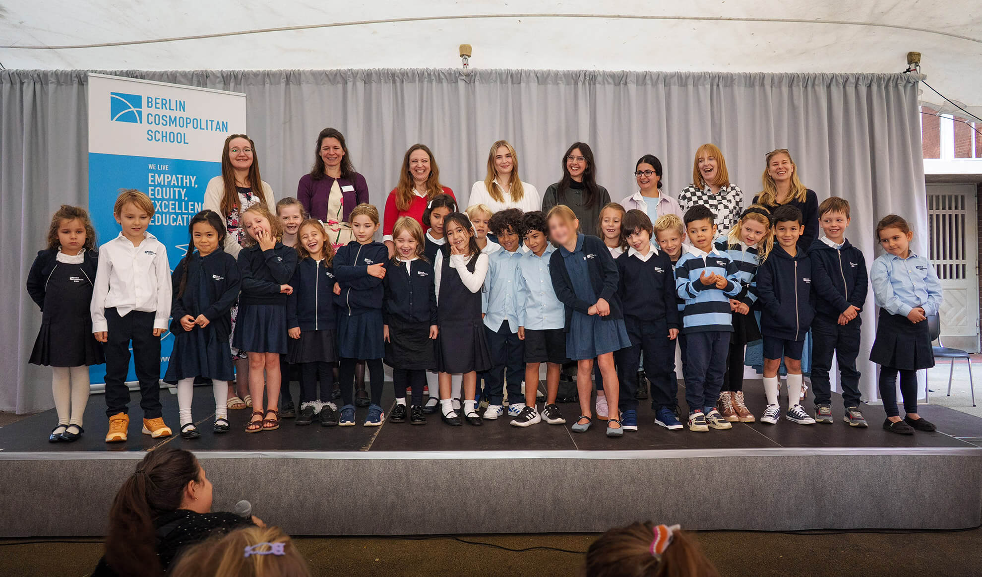 Cosmopolitanschool Berlin Eine Gruppe junger Kinder und mehrere Erwachsene stehen auf einer Bühne und lächeln für ein Foto bei der Einschulungsfeier der Berlin Cosmopolitan School - ein besonderer Moment. Auf der linken Seite ist ein Banner mit dem Namen der Schule zu sehen, dahinter ein Vorhang. loading=