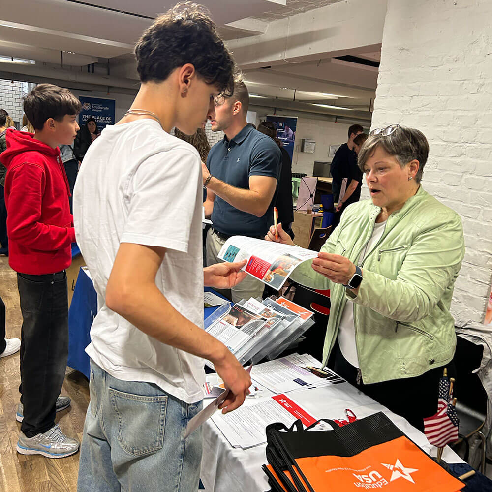 Cosmopolitanschool Berlin Beim International College Day überreicht eine Frau an einem BCS-Stand einem jungen Mann eine Broschüre. Andere Menschen unterhalten sich an den Tischen in der Nähe, nehmen Informationsmaterial und Tragetaschen in die Hand und bringen die Schüler mit neuen Möglichkeiten in Kontakt.