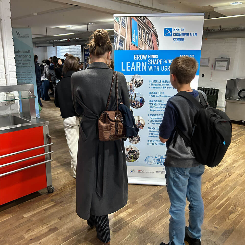 Cosmopolitanschool Berlin Zwei Personen mit Rucksäcken stehen vor einem Banner der Berlin Cosmopolitan School (BCS) während des International College Day, der Schüler in einer Halle mit Holzfußboden miteinander verbindet, während sich im Hintergrund andere versammeln.