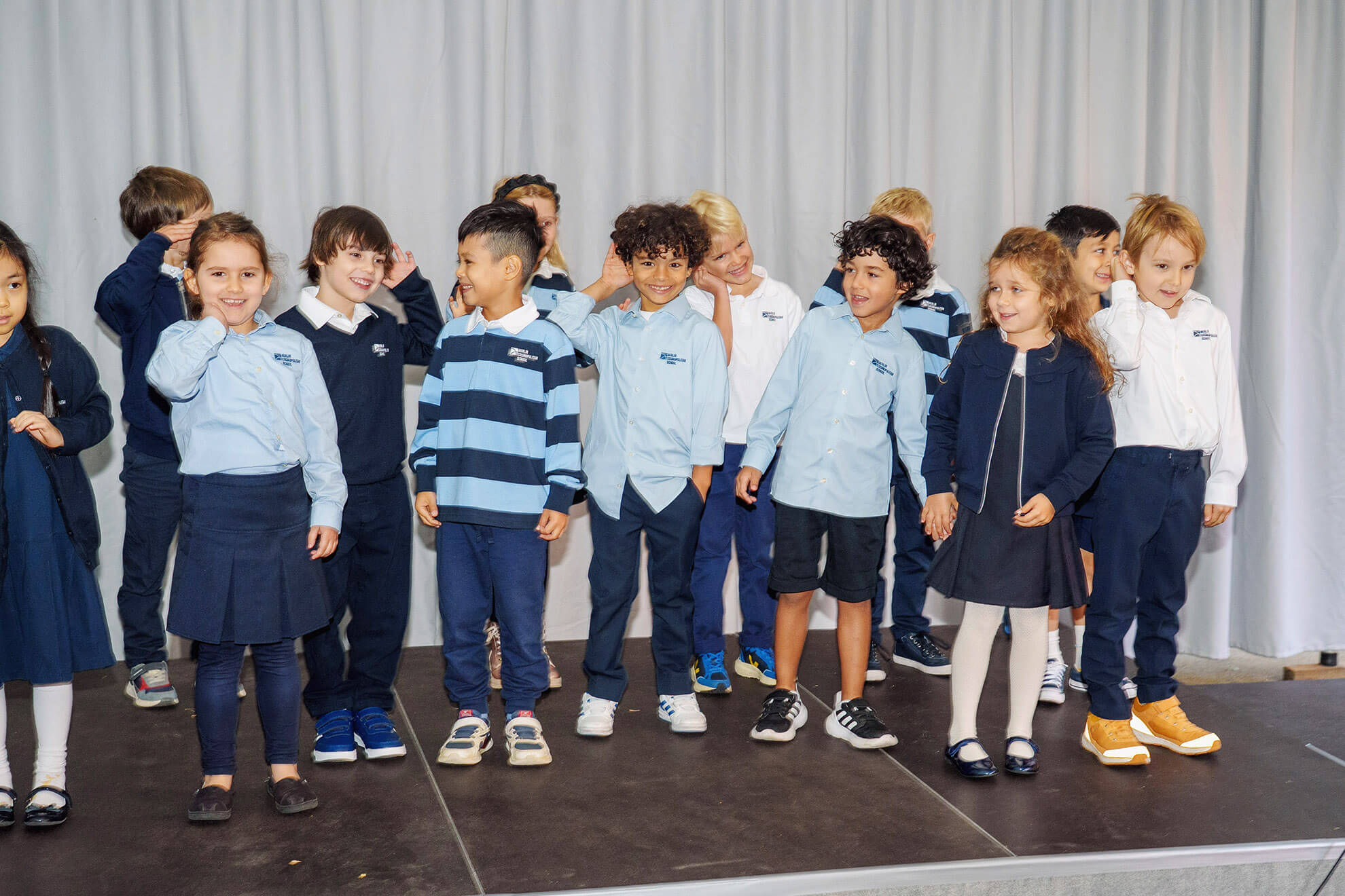 Cosmopolitanschool Berlin Eine Gruppe junger Kinder in blau-weißen Uniformen steht bei der Einschulungsfeier der Berlin Cosmopolitan School auf der Bühne, lächelt und interagiert miteinander vor einem hellgrauen Vorhang. loading=