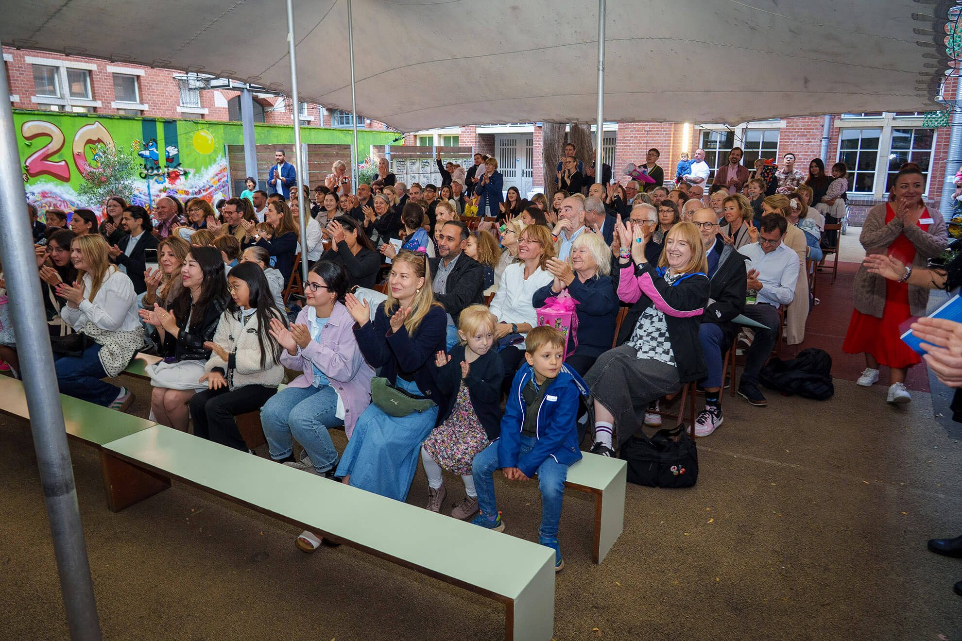 Cosmopolitanschool Berlin Eine große Gruppe von Erwachsenen und Kindern sitzt dicht beieinander unter einem Zelt, klatscht und lächelt, während sie die Einschulungsfeier der Berlin Cosmopolitan School verfolgen - ein besonderer Moment. Im Hintergrund sind bunte Dekorationen und ein Wandgemälde mit der Zahl 20 zu sehen. loading=