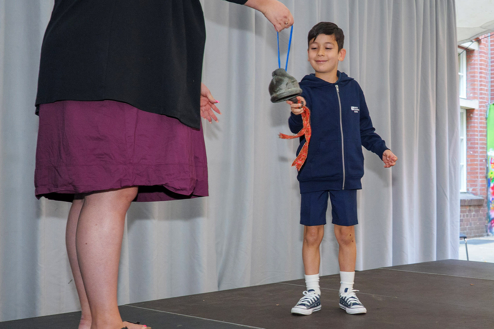 Cosmopolitanschool Berlin Ein kleiner Junge in einem marineblauen Outfit lächelt, als er während der Einschulungsfeier in der Berlin Cosmopolitan School eine Glocke an einem Band von einem Erwachsenen erhält - ein besonderer Moment auf der Bühne mit grauen Vorhängen im Hintergrund.