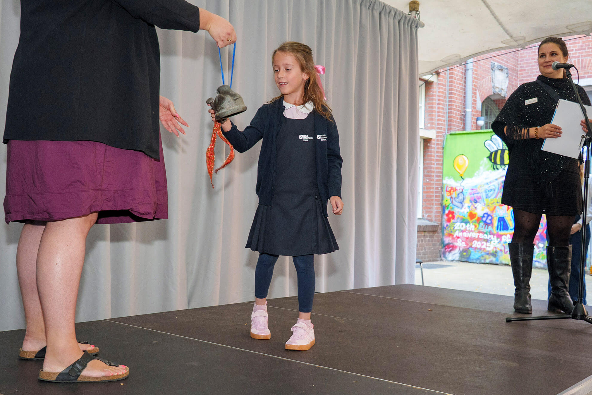 Cosmopolitanschool Berlin Auf der Einschulungsfeier der Berliner Cosmopolitan School lächelt ein junges Mädchen in einem marineblauen Kleid, als sie von einem Erwachsenen auf der Bühne eine Glocke entgegennimmt - ein besonderer Moment, mit einem farbenfrohen Wandgemälde und einer anderen Frau in der Nähe, die ein Mikrofon hält.