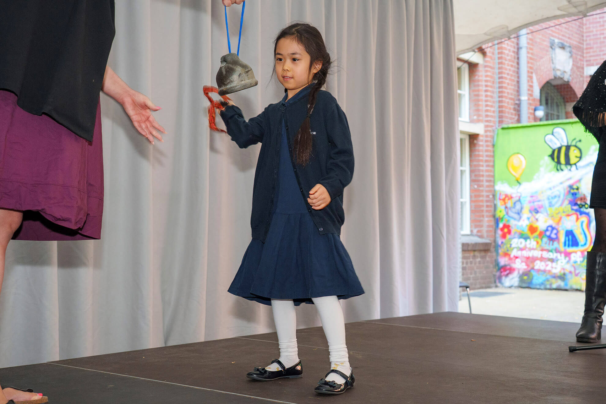 Cosmopolitanschool Berlin Bei der Einschulungsfeier der Berliner Cosmopolitan School steht ein junges Mädchen in einem marineblauen Kleid und einer weißen Strumpfhose auf der Bühne und hält eine Glocke in der Hand - und fängt einen besonderen Moment ein, während zwei Erwachsene vor einem farbenfrohen Wandhintergrund nach ihr greifen.
