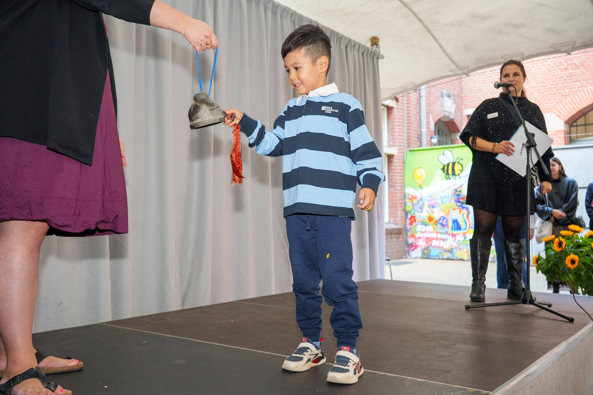 Cosmopolitanschool Berlin Bei der Einschulungsfeier der Berliner Cosmopolitan School läutet ein Junge in einem gestreiften Hemd und Turnschuhen eine Glocke, die ihm ein Erwachsener auf der Bühne reicht, während im Hintergrund eine lächelnde Frau mit einem Mikrofon steht.