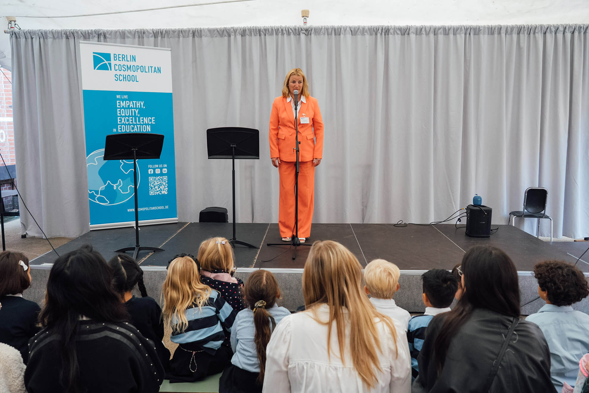 Cosmopolitanschool Berlin Eine Frau in einem leuchtend orangefarbenen Anzug spricht auf der Bühne der Einschulungsfeier der Berlin Cosmopolitan School vor einem Publikum aus Kindern und Erwachsenen. Neben ihr steht ein Schulbanner, graue Vorhänge bilden die Kulisse für diesen besonderen Moment. loading=