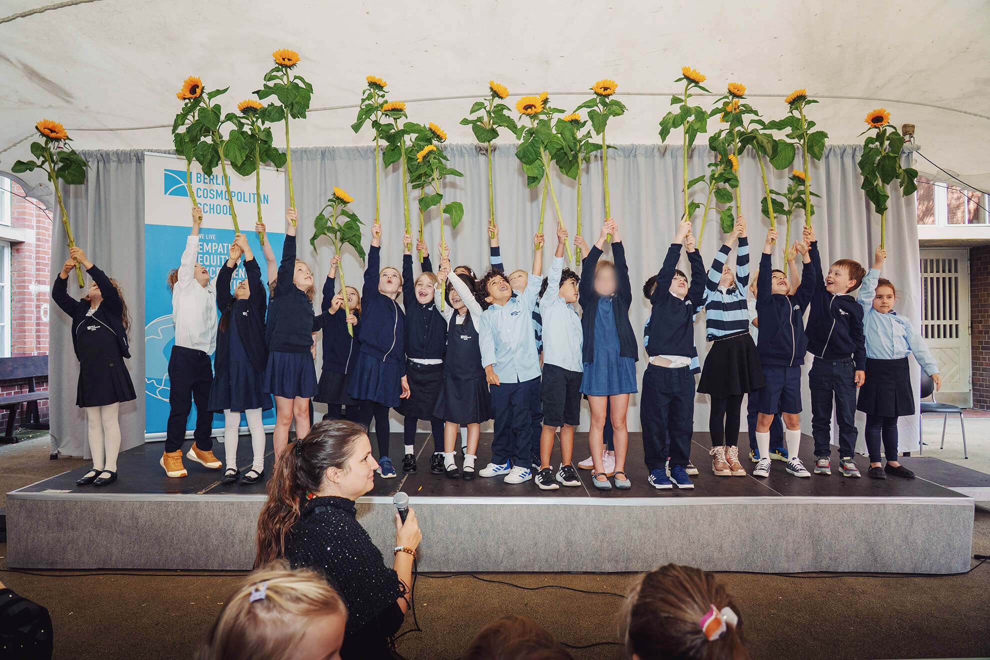 Cosmopolitanschool Berlin Bei der Einschulungsfeier der Berlin Cosmopolitan School steht eine Gruppe von Kindern in Schuluniformen auf der Bühne unter einem Zelt und hält jeweils eine große Sonnenblume über ihren Köpfen - und fängt damit diesen besonderen Moment für alle Anwesenden ein. loading=