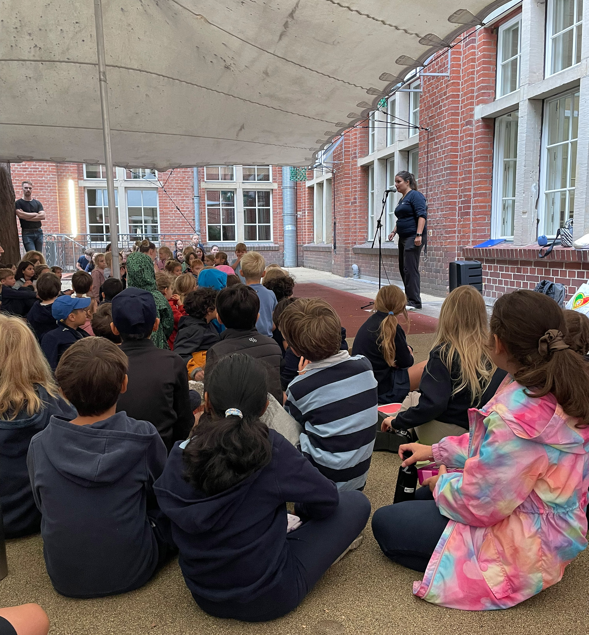 Cosmopolitanschool Berlin Eine Gruppe von Kindern sitzt auf dem Boden unter einem großen Vordach und beobachtet einen Mann, der auf einer kleinen Freiluftbühne in der Nähe eines gemauerten Schulgebäudes spricht. Einige Erwachsene stehen in der Nähe. Es ist wahrscheinlich eine Grundschulversammlung für einen fröhlichen Start ins Schuljahr. loading=