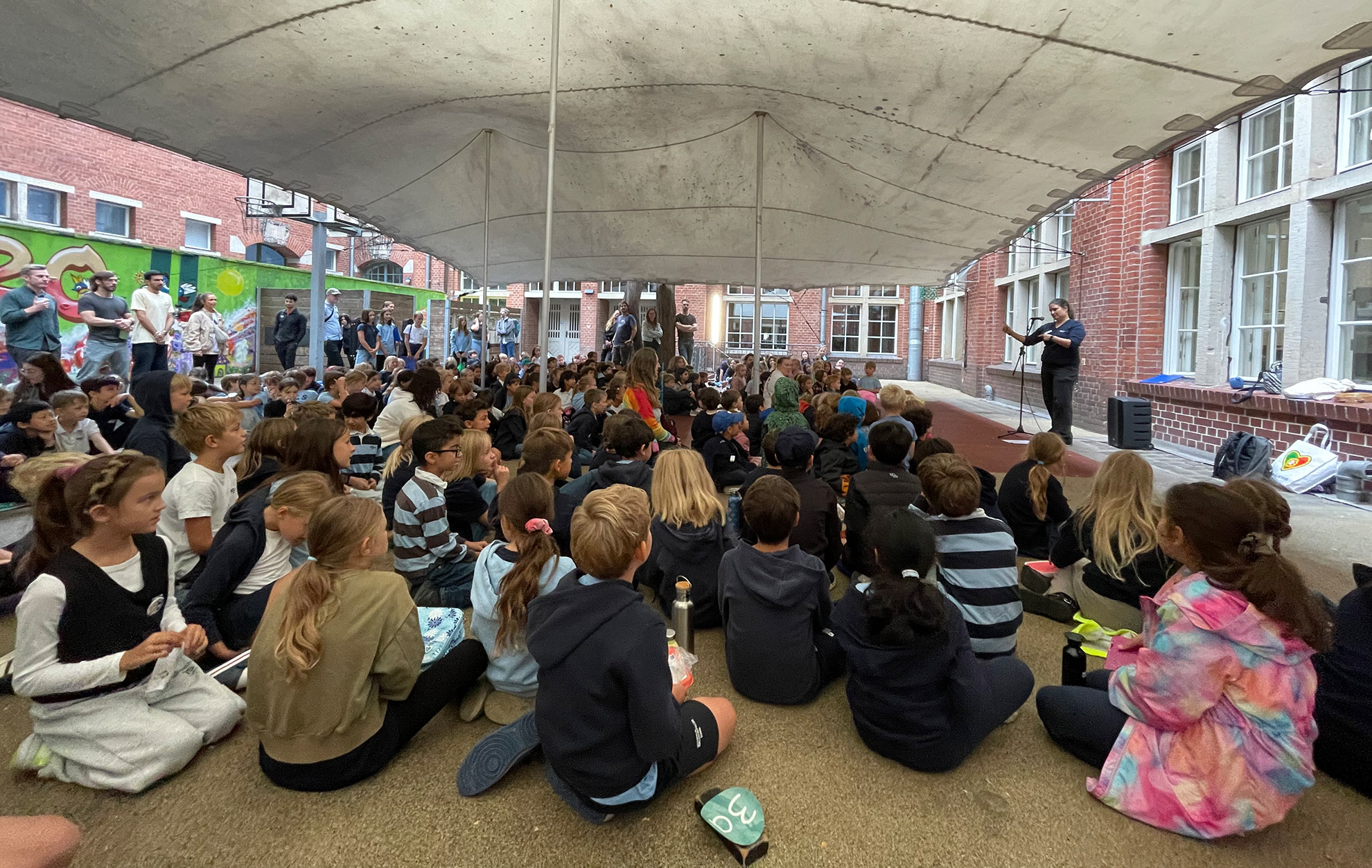 Cosmopolitanschool Berlin Eine große Gruppe von Kindern sitzt auf dem Boden unter einem weißen Baldachin und beobachtet aufmerksam eine Person, die auf einer kleinen Bühne vor einem Backsteingebäude spricht. Diese lebendige Grundschulversammlung markiert einen fröhlichen Start in das Schuljahr. loading=