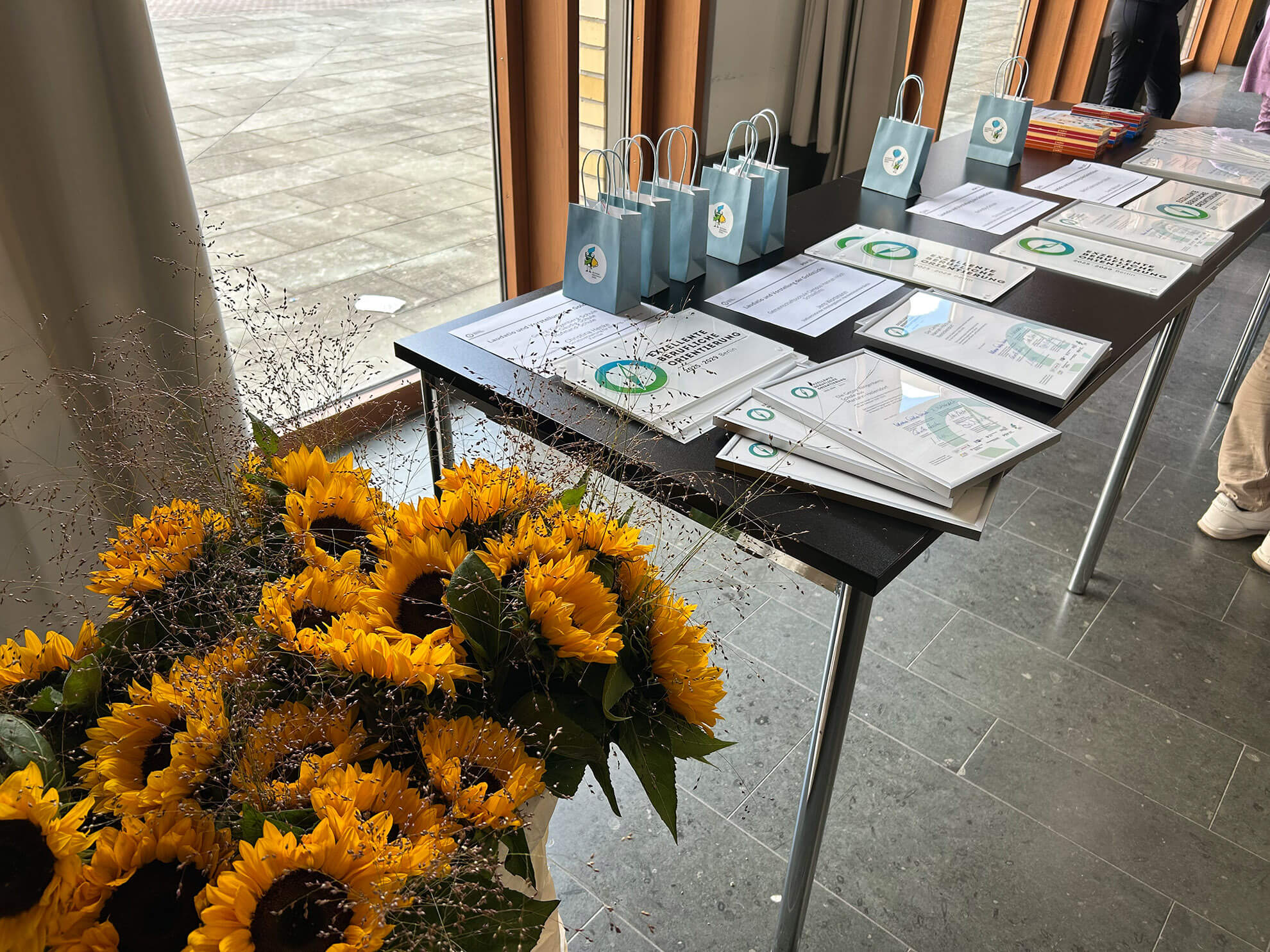 Cosmopolitanschool Berlin Auf einem Tisch liegen Zertifikate, Geschenktüten und Mappen - einige mit dem Qualitätssiegel der Berlin Cosmopolitan School - neben einem großen Strauß Sonnenblumen. Die Umgebung scheint ein Innenraum zu sein, in der Nähe von Fenstern mit Blick auf einen gepflasterten Außenbereich.