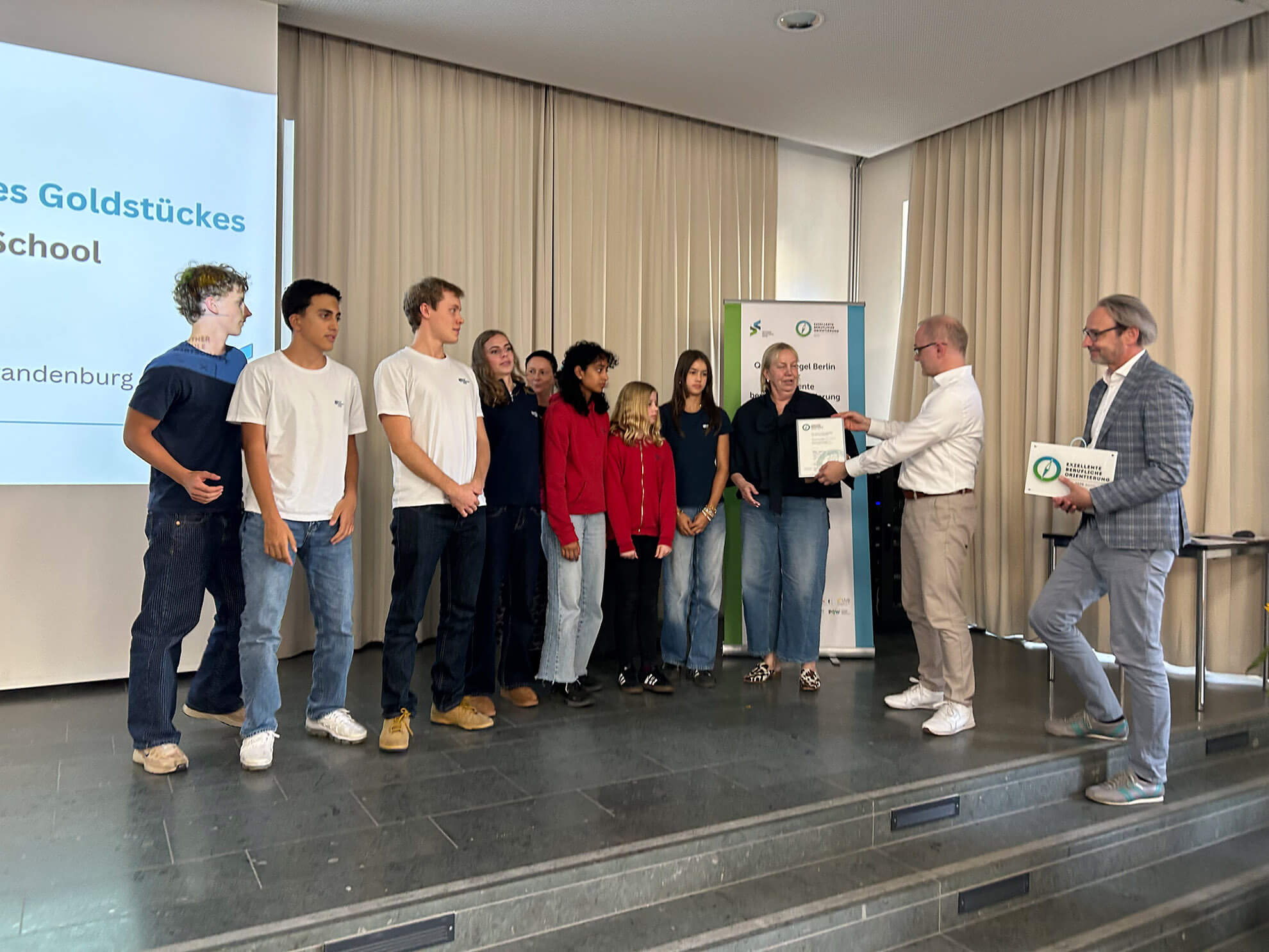 Cosmopolitanschool Berlin Eine Gruppe von Schülern der Berlin Cosmopolitan School steht auf der Bühne und erhält von zwei Männern in Anzügen das Qualitätssiegel für exzellente Berufsorientierung. Einer überreicht eine Urkunde, während die anderen zusehen. Im Hintergrund sind eine Präsentationsfolie und ein Banner zu sehen. loading=