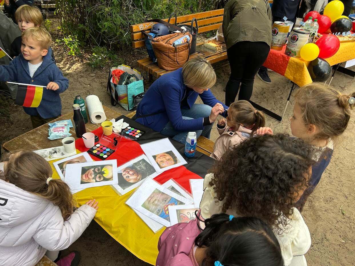 Cosmopolitanschool Berlin Kinder versammeln sich um einen mit einer deutschen Flagge, Farben und Fotos geschmückten Tisch während der Veranstaltung zum Internationalen Tag der Berlin Cosmopolitan School. Ein Erwachsener malt das Gesicht eines Mädchens, während andere zusehen. Luftballons und Zubehör sind in der Nähe im Kindergartenbereich zu sehen.