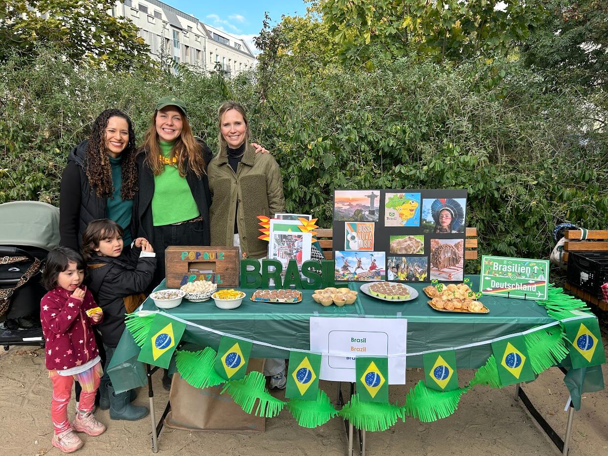 Cosmopolitanschool Berlin Vier Frauen und zwei Kinder stehen hinter einem Tisch, der mit brasilianischen Flaggen, Lebensmitteln und farbenfrohen Schautafeln über Brasilien geschmückt ist, beim Internationalen Tag der Berlin Cosmopolitan School. Sie befinden sich im Freien, umgeben von Grünflächen und Gebäuden im Hintergrund.