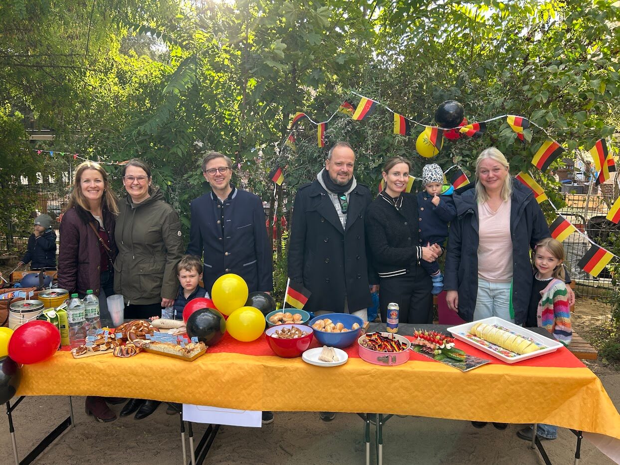 Cosmopolitanschool Berlin Eine Gruppe von Erwachsenen und Kindern aus dem Kindergarten der Berlin Cosmopolitan School steht hinter einem Tisch, der mit Snacks, Getränken und der deutschen Flagge geschmückt ist, bei einer Veranstaltung zum Internationalen Tag im Freien. Im Hintergrund sind Bäume und Festtagsbanner zu sehen.