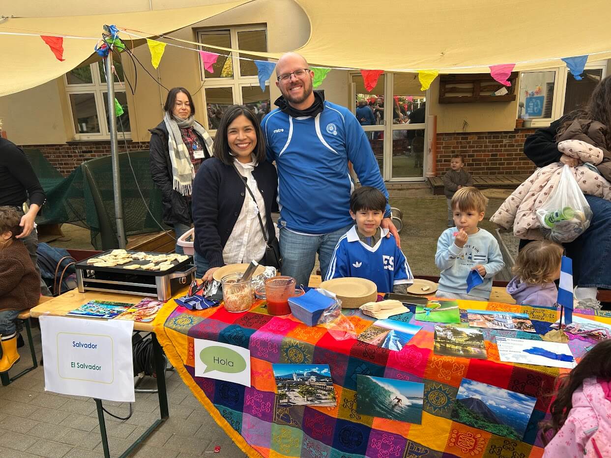 Cosmopolitanschool Berlin Ein lächelnder Mann und eine lächelnde Frau stehen hinter einem farbenfrohen Tisch mit salvadorianischen Speisen und Fotos beim Internationalen Tag der Berlin Cosmopolitan School, zu dem auch Kindergartenkinder kommen. Auf dem Tisch steht ein "Hola"-Schild, darüber hängen festliche Wimpel im Freien.