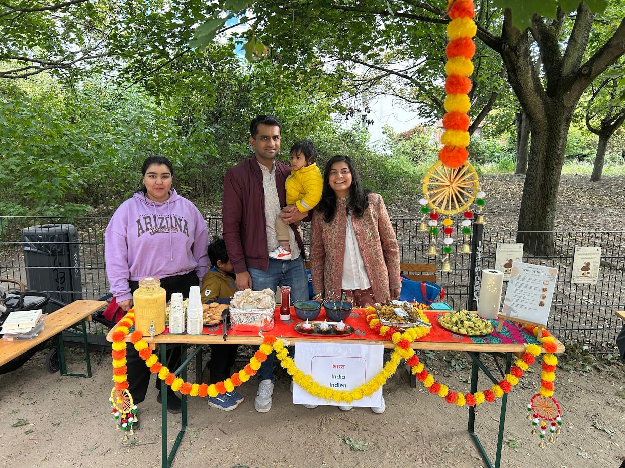 Cosmopolitanschool Berlin Vier Personen, darunter ein Kind aus dem Kindergarten der Berlin Cosmopolitan School, stehen hinter einem geschmückten Tisch mit indischen Speisen und Getränken zum Internationalen Tag. Ringelblumengirlanden und ein Schild mit der Aufschrift "Indien Indien" schmücken die Szene in einem Park.