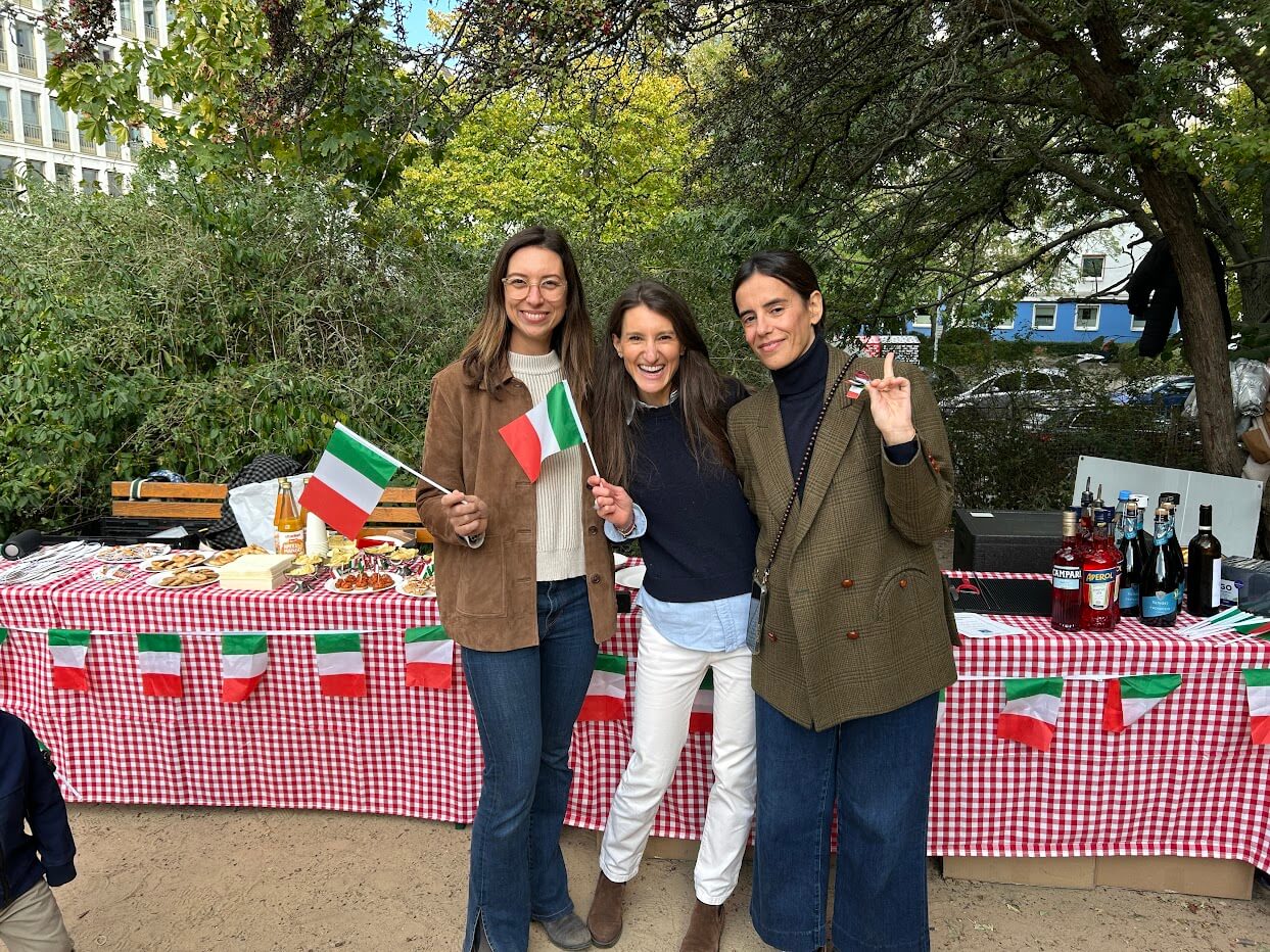 Cosmopolitanschool Berlin Drei Frauen stehen lächelnd vor einem mit italienischen Flaggen und rot karierten Tischdecken geschmückten Picknicktisch beim Internationalen Tag der Berlin Cosmopolitan School. Zwei Frauen halten kleine italienische Flaggen, auf denen Speisen und Getränke für die Kindergartenveranstaltung im Freien ausgestellt sind.