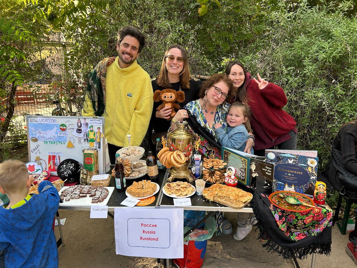 Cosmopolitanschool Berlin Fünf lächelnde Menschen, darunter zwei Kinder aus dem Kindergarten der Berlin Cosmopolitan School, stehen hinter einem Tisch mit russischen Lebensmitteln und Kulturgütern zum Internationalen Tag. Auf dem Tisch stehen ein Samowar, Gebäck, dekorative Tücher und Grünzeug im Hintergrund.