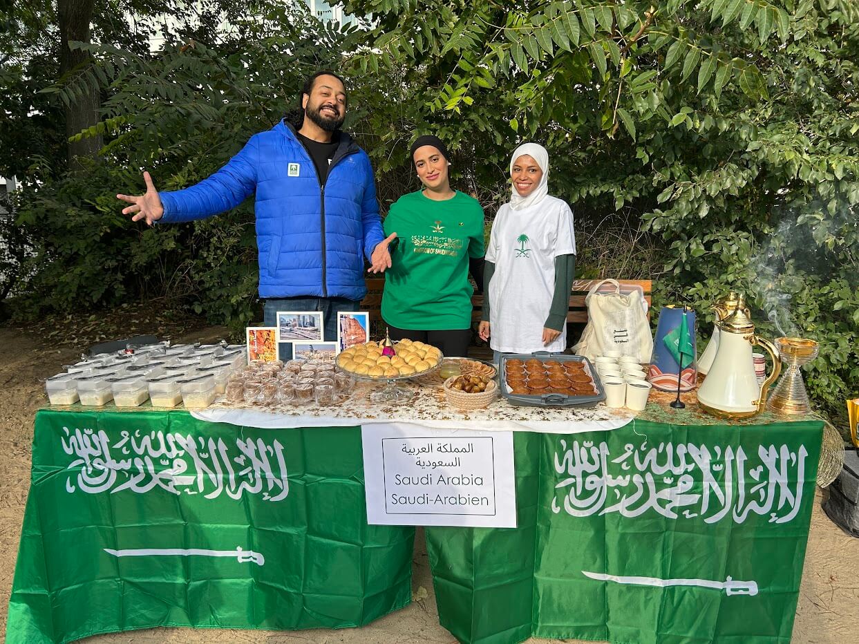 Cosmopolitanschool Berlin Beim Internationalen Tag der Berlin Cosmopolitan School stehen drei Personen hinter einem mit saudi-arabischen Flaggen bedeckten Tisch und bieten traditionelle Speisen, Desserts, saudischen Kaffee und Tee an. Auf einem Schild steht "Saudi-Arabien" auf Arabisch, Englisch und Deutsch.