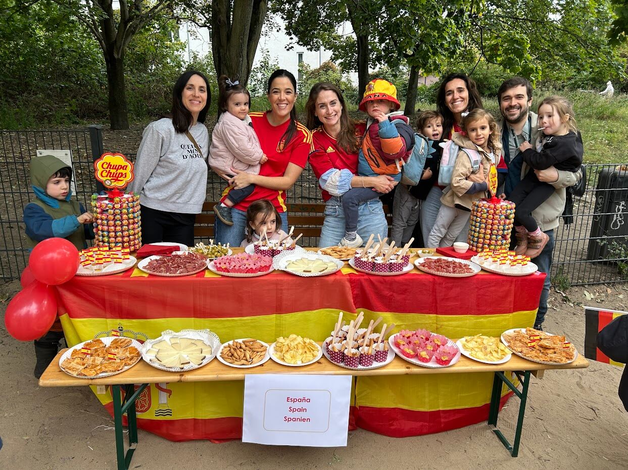 Cosmopolitanschool Berlin Eine Gruppe von Erwachsenen und Kindern aus dem Kindergarten der Berlin Cosmopolitan School steht hinter einem mit einer spanischen Flagge bedeckten Tisch, auf dem spanische Speisen und Leckereien für den Internationalen Tag ausgestellt sind. Luftballons und Chupa Chups Lutscher schmücken die festliche Szene.