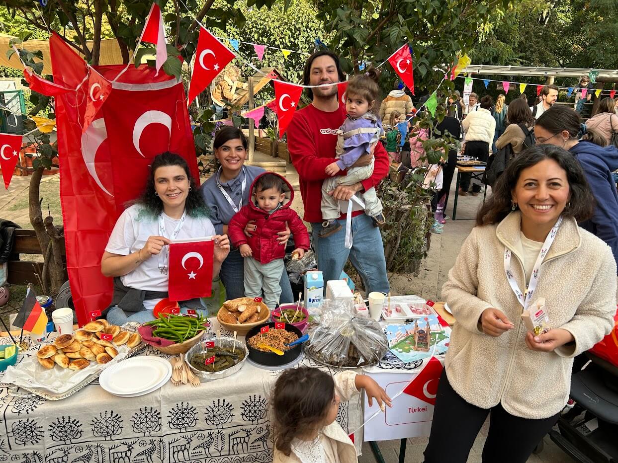 Cosmopolitanschool Berlin Eine Gruppe lächelnder Menschen, darunter auch Kinder des Kindergartens der Berlin Cosmopolitan School, versammeln sich an einem türkischen Stand mit Fahnen, Gebäck und Süßigkeiten während des Internationalen Tages, mit bunten Wimpeln und anderen im Hintergrund.