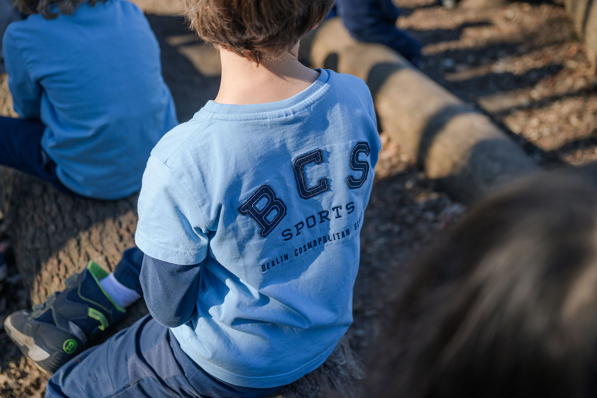 Cosmopolitanschool Berlin Ein Kind sitzt auf einem Baumstamm im Freien und trägt ein blaues T-Shirt mit dem Aufdruck BCS SPORTS und Berlin Cosmopolitan School auf dem Rücken. Andere Kinder, die möglicherweise an einem Cosmo-Swap teilnehmen, sind in der Nähe und tragen ähnliche blaue Kleidung. loading=