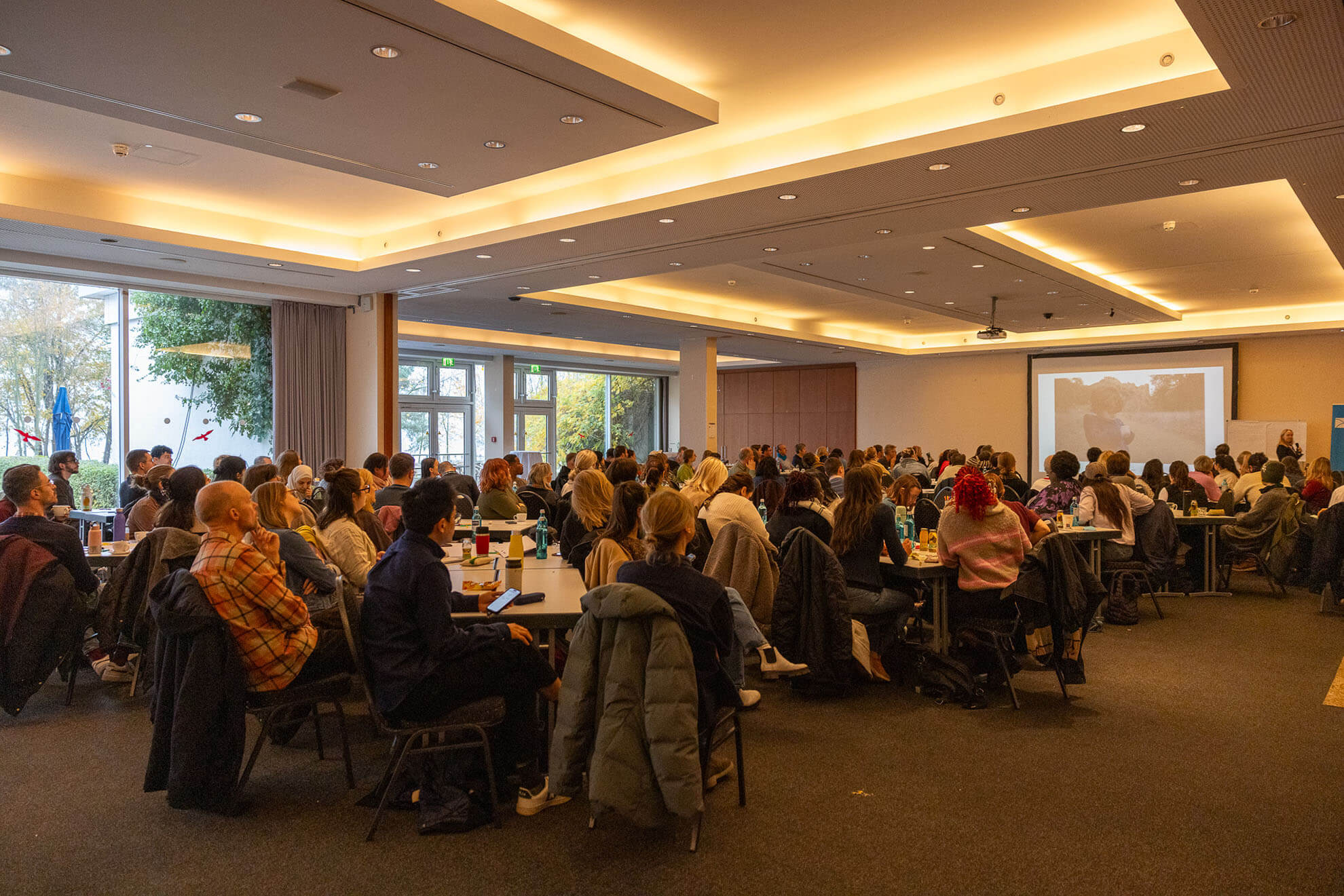 Cosmopolitanschool Berlin Eine große Gruppe von Menschen sitzt an runden Tischen in einem geräumigen, gut beleuchteten Konferenzraum und verfolgt aufmerksam die Präsentation von Karth Murdoch, die auf eine Leinwand an der Stirnseite projiziert wird. An den Stühlen hängen Mäntel, und aus den Fenstern kann man die Bäume draußen sehen.