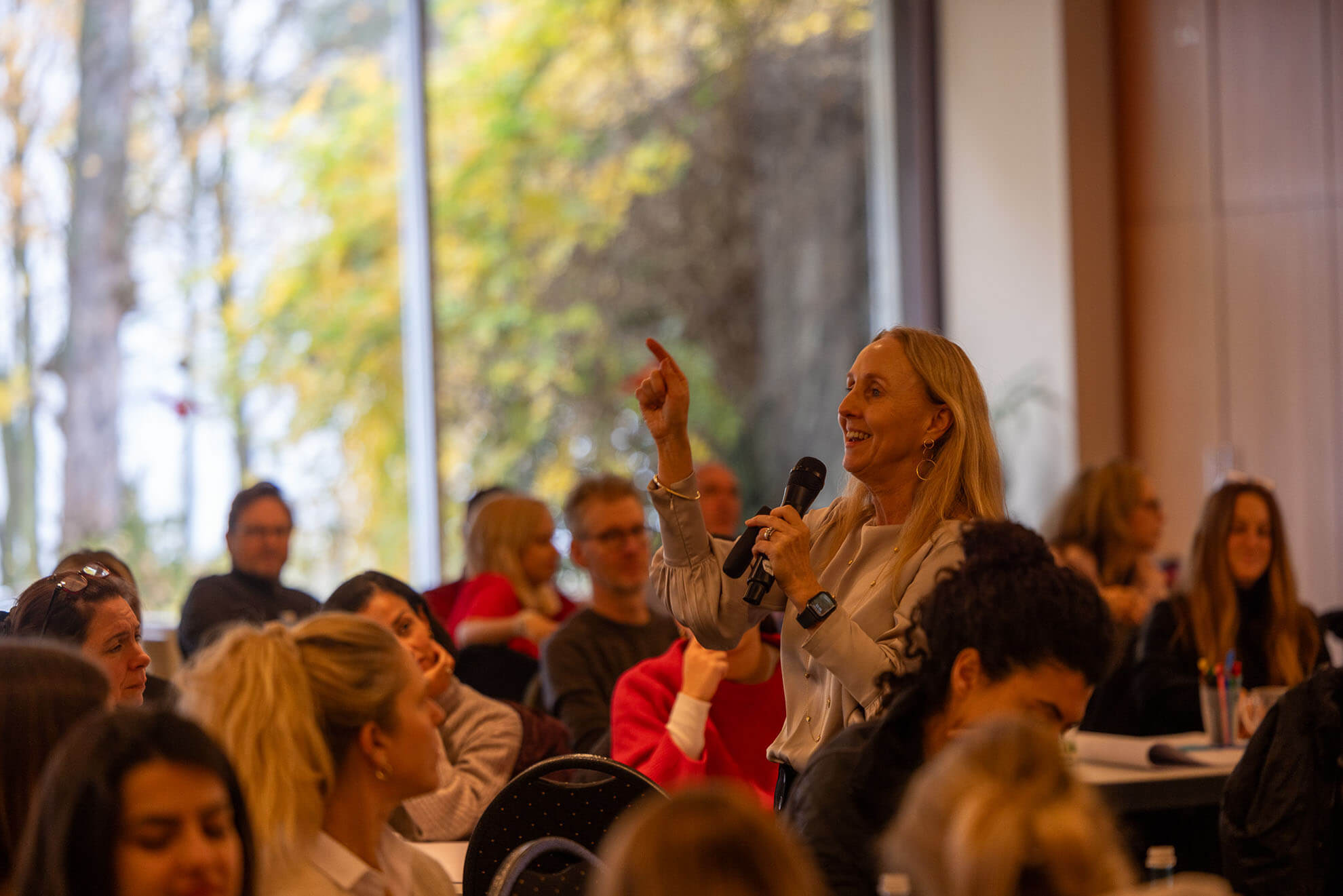 Cosmopolitanschool Berlin Karth Murdoch steht mit einem Mikrofon in der Hand und gestikuliert, während er zu einem sitzenden Publikum in einem hellen, modernen Raum mit großen Fenstern und Blick auf die Bäume draußen spricht. Das Publikum hört aufmerksam zu.
