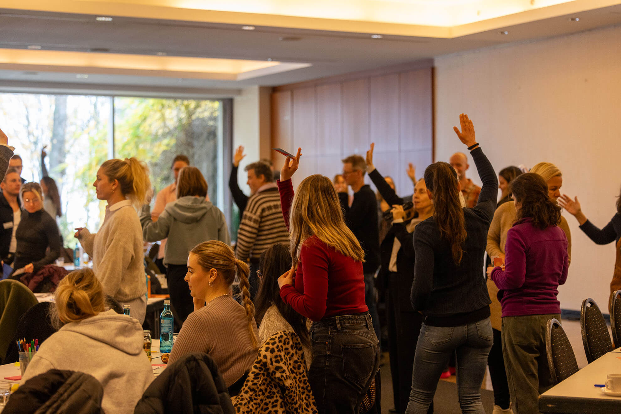 Cosmopolitanschool Berlin Eine Gruppe von Personen in einem Konferenzraum steht und hebt die Hände, um an einer interaktiven Sitzung unter der Leitung von Karth Murdoch teilzunehmen. Einige sprechen miteinander, während andere nach vorne schauen. Tische mit Material sind zu sehen. loading=