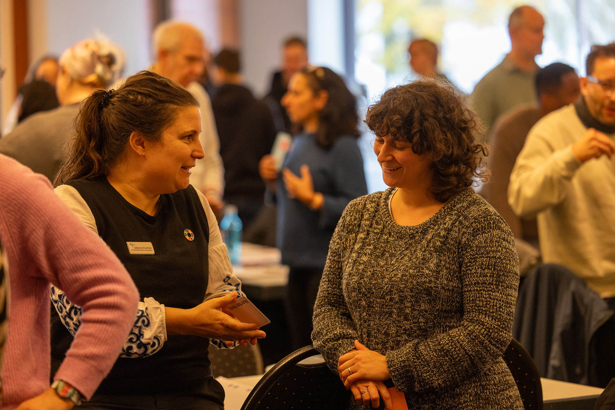 Cosmopolitanschool Berlin Zwei Frauen stehen und lächeln, während sie sich in einem belebten, gut beleuchteten Raum mit vielen Menschen unterhalten. Eine Frau, die möglicherweise Neuigkeiten über Karth Murdoch mitteilt, hält ein Telefon und gestikuliert, während andere im Hintergrund ihre Gespräche fortsetzen.