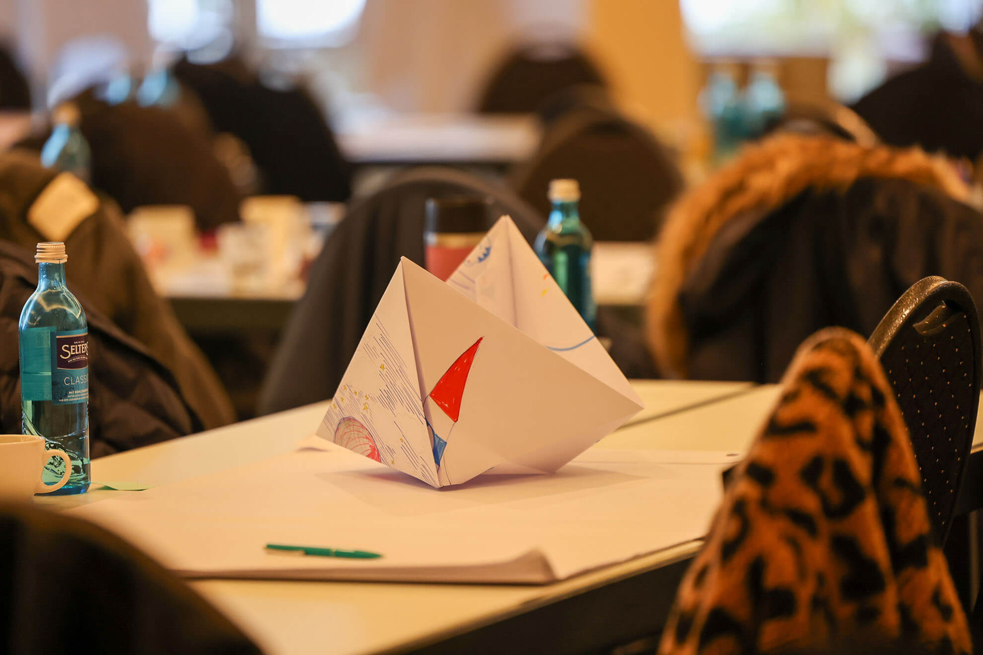 Cosmopolitanschool Berlin Ein gefaltetes Papiersegelboot mit bunten Zeichnungen liegt auf einem Tisch in einem Konferenzraum, umgeben von Wasserflaschen, Stiften, Mänteln, die über Stühle drapiert sind, und unscharfen Hintergrundteilnehmern an der Karth Murdoch Veranstaltung.