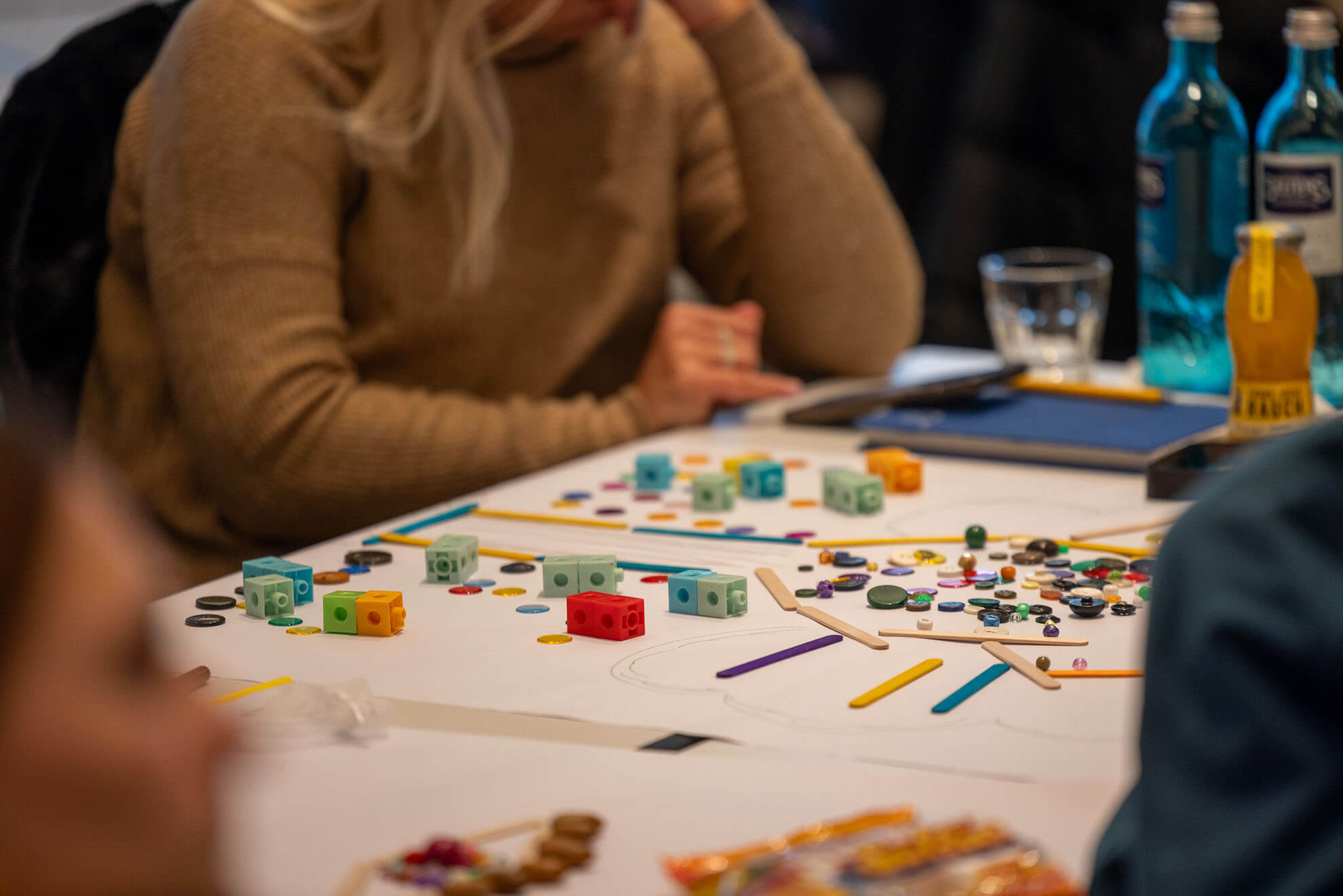 Cosmopolitanschool Berlin Karth Murdoch sitzt an einem Tisch in einem hellbraunen Pullover, umgeben von bunten Würfeln, Knöpfen, Stöcken und anderen Spiel- oder Bastelteilen, die auf eine kreative oder gemeinschaftliche Workshop-Aktivität hindeuten. Im Hintergrund stehen Wasserflaschen.