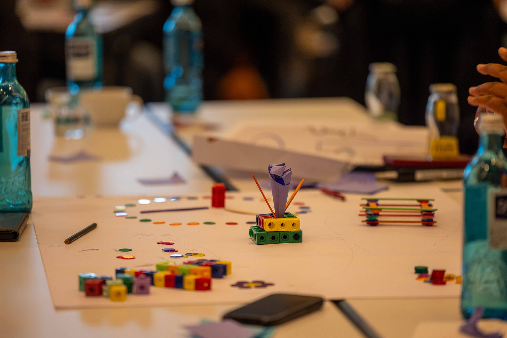Cosmopolitanschool Berlin Ein Tisch mit bunten Bauklötzen, Bastelmaterial, Papier, Stiften und Wasserflaschen für eine von Karth Murdoch inspirierte kreative Gruppenaktivität. Die Hände sind bei der Arbeit an einem Projekt zu sehen.
