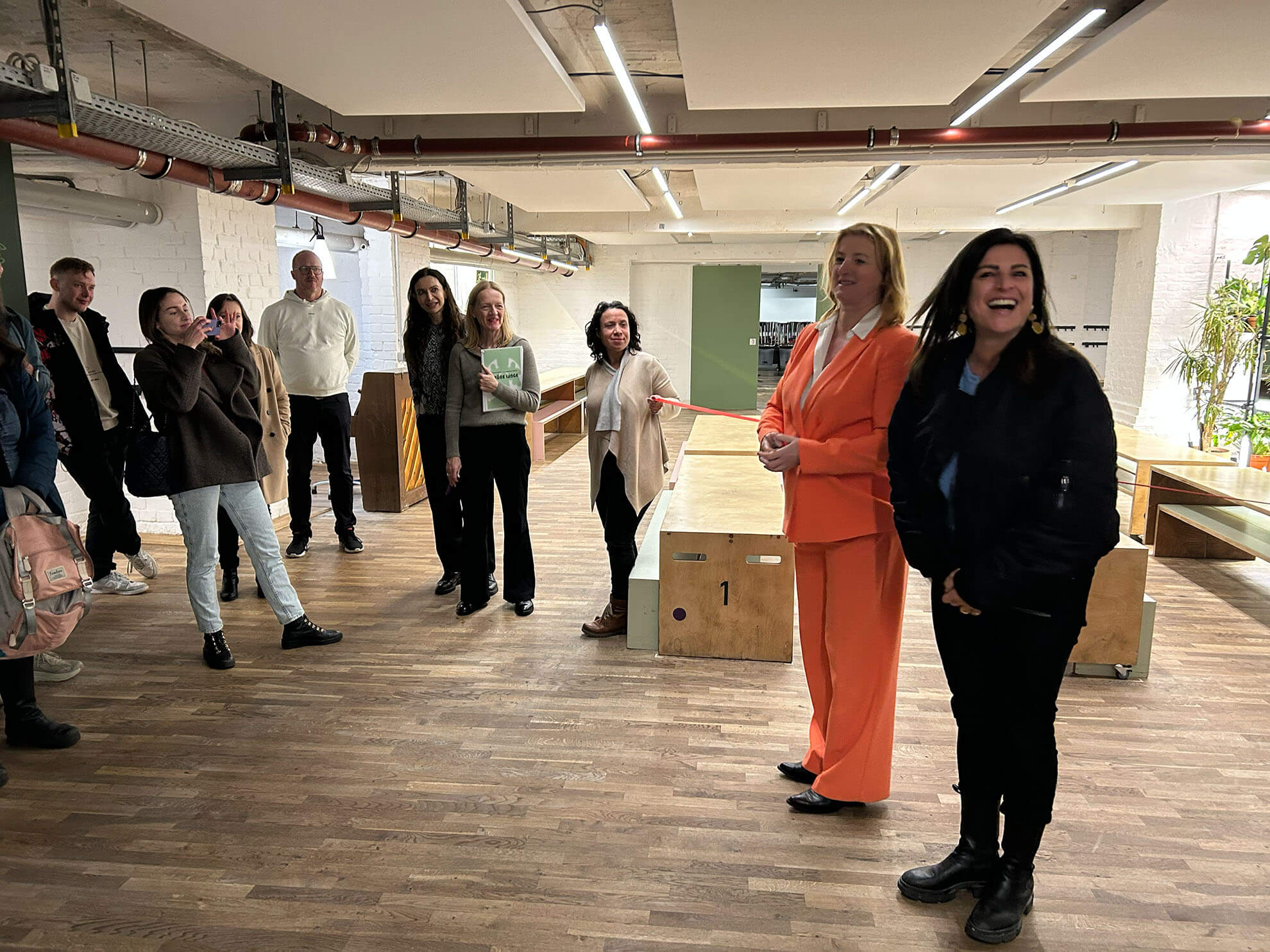 Cosmopolitanschool Berlin Eine Gruppe von Menschen versammelt sich in einem Büroraum, um im Geiste der Gemeinschaft zu feiern. In der Mitte schneidet eine Person in einem orangefarbenen Anzug ein rotes Band durch, während andere lächelnd zusehen und die feierliche Eröffnung der neuen BCS-Cafeteria feiern.