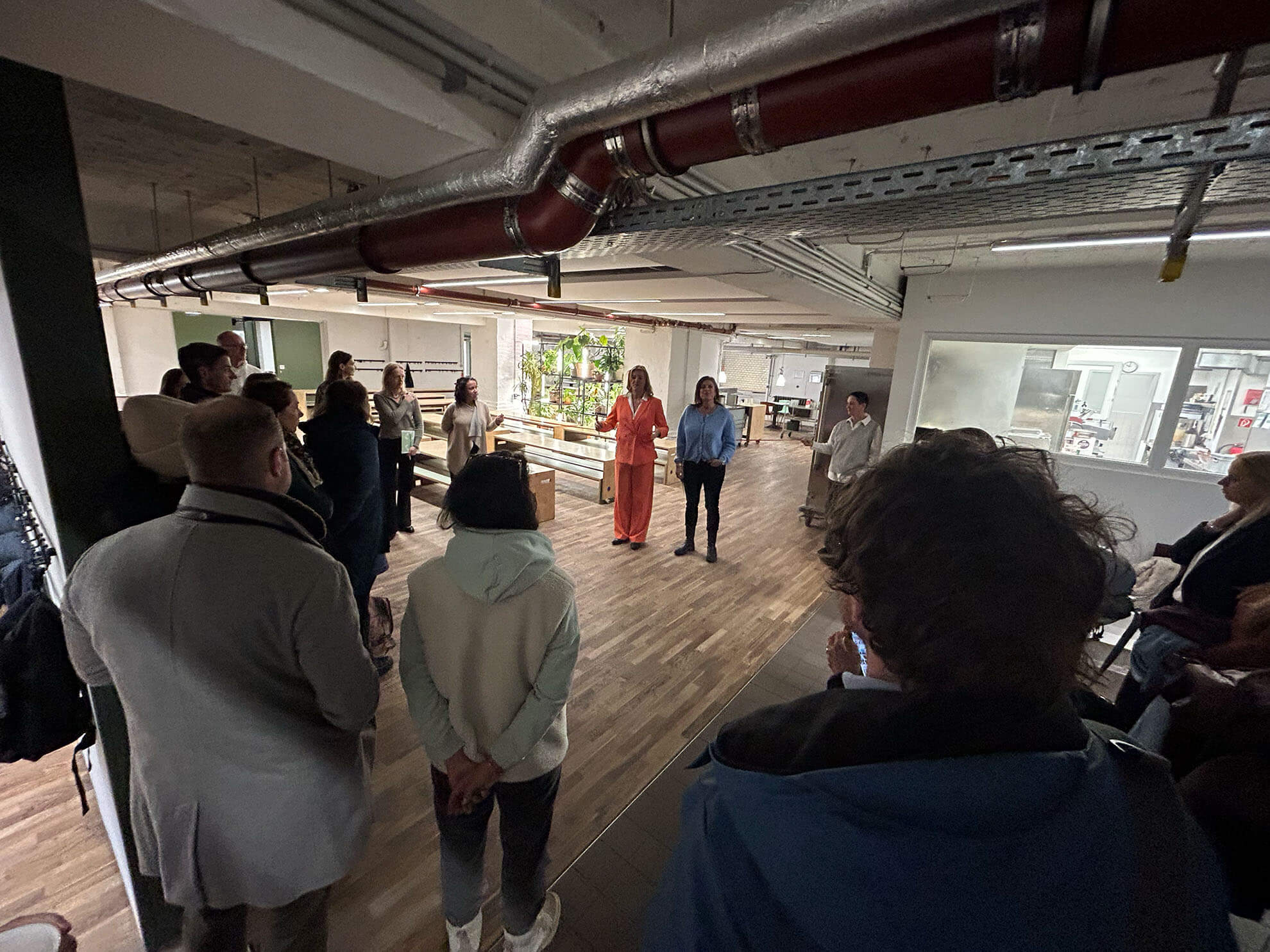 Cosmopolitanschool Berlin Eine Gruppe von Menschen steht in einem modernen Büroraum und pflegt die Gemeinschaft, während sie zwei Lautsprechern in der Mitte zuhört. Der Raum ist mit Holzböden, freiliegenden Rohren an der Decke und verglasten Büros im Hintergrund ausgestattet.