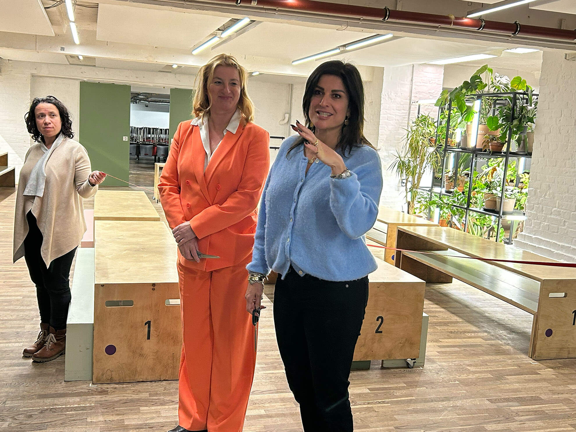 Cosmopolitanschool Berlin Drei Frauen stehen in einer modernen Cafeteria mit Holzbänken und Grünpflanzen. Gekleidet in einen orangefarbenen Anzug, eine hellblaue Jacke und einen beigen Pullover, verkörpern sie ein Gefühl von Gemeinschaft, während sie sich angeregt unterhalten.