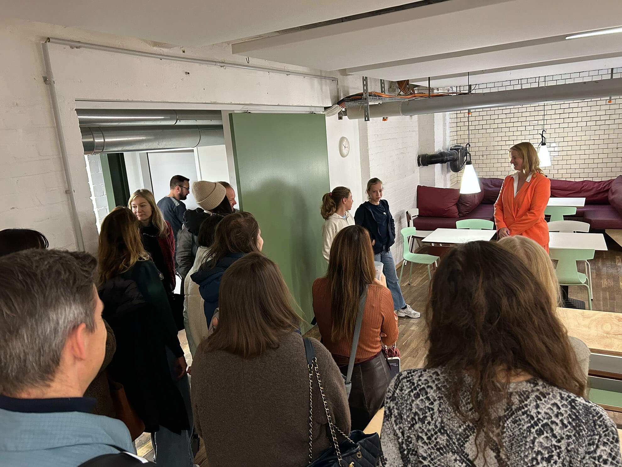 Cosmopolitanschool Berlin Eine Gruppe von Menschen steht in einer Cafeteria und hört einer Frau in einem orangefarbenen Kleid zu. Der Raum, der die Gemeinschaft fördert, ist mit grünen Türen, weißen Backsteinwänden, Tischen, Stühlen und einem roten Sofa im Hintergrund ausgestattet.