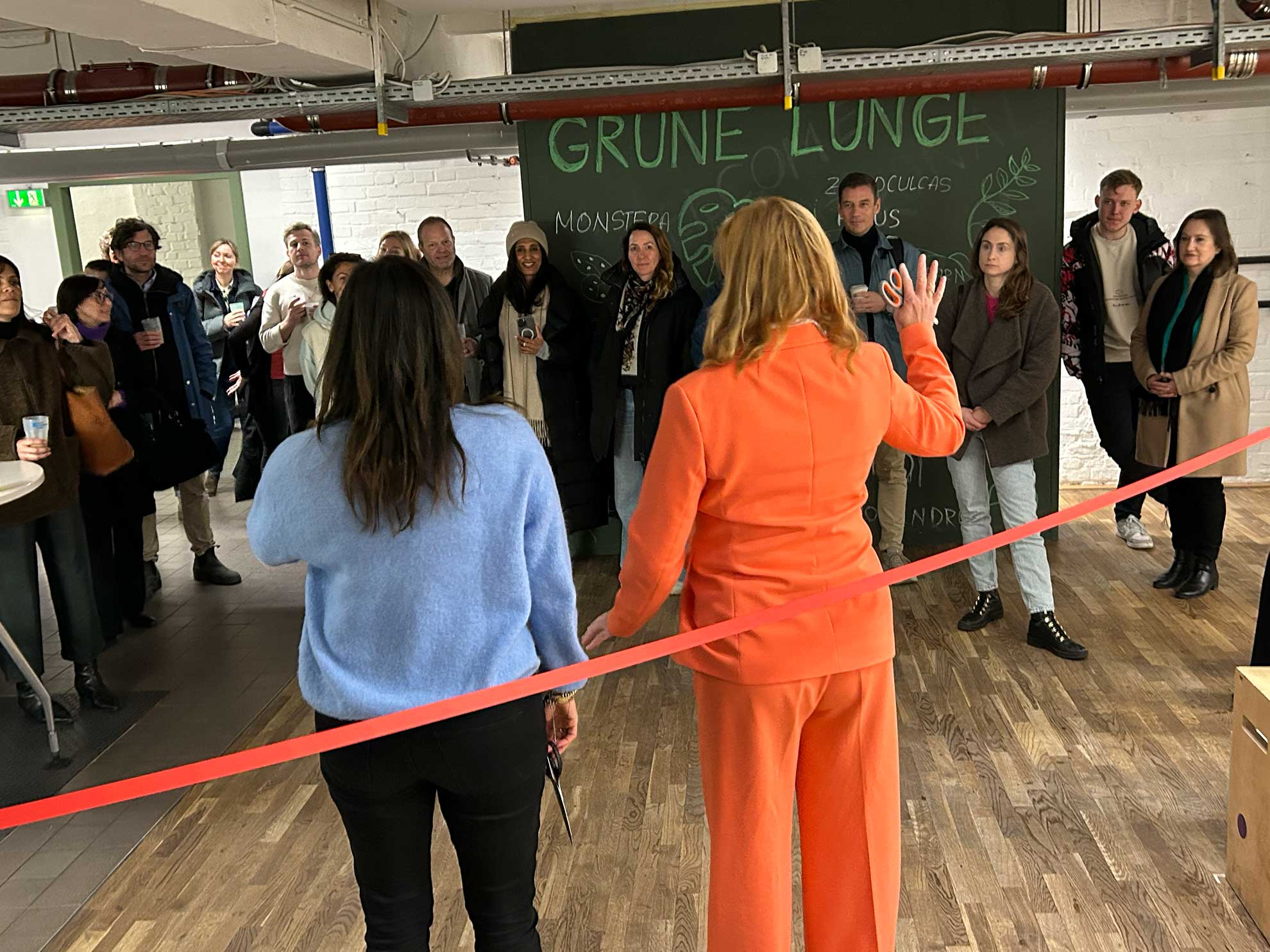 Cosmopolitanschool Berlin Eine Gruppe von Menschen beobachtet, wie zwei Frauen, eine im orangefarbenen Anzug und eine im blauen Pullover, bei einer BCS-Indoor-Veranstaltung zur Förderung der Gemeinschaft vor einer roten Schleife stehen, auf der mit Kreide die Aufschrift GRUNE LUNGE in der Nähe der Cafeteria zu lesen ist.