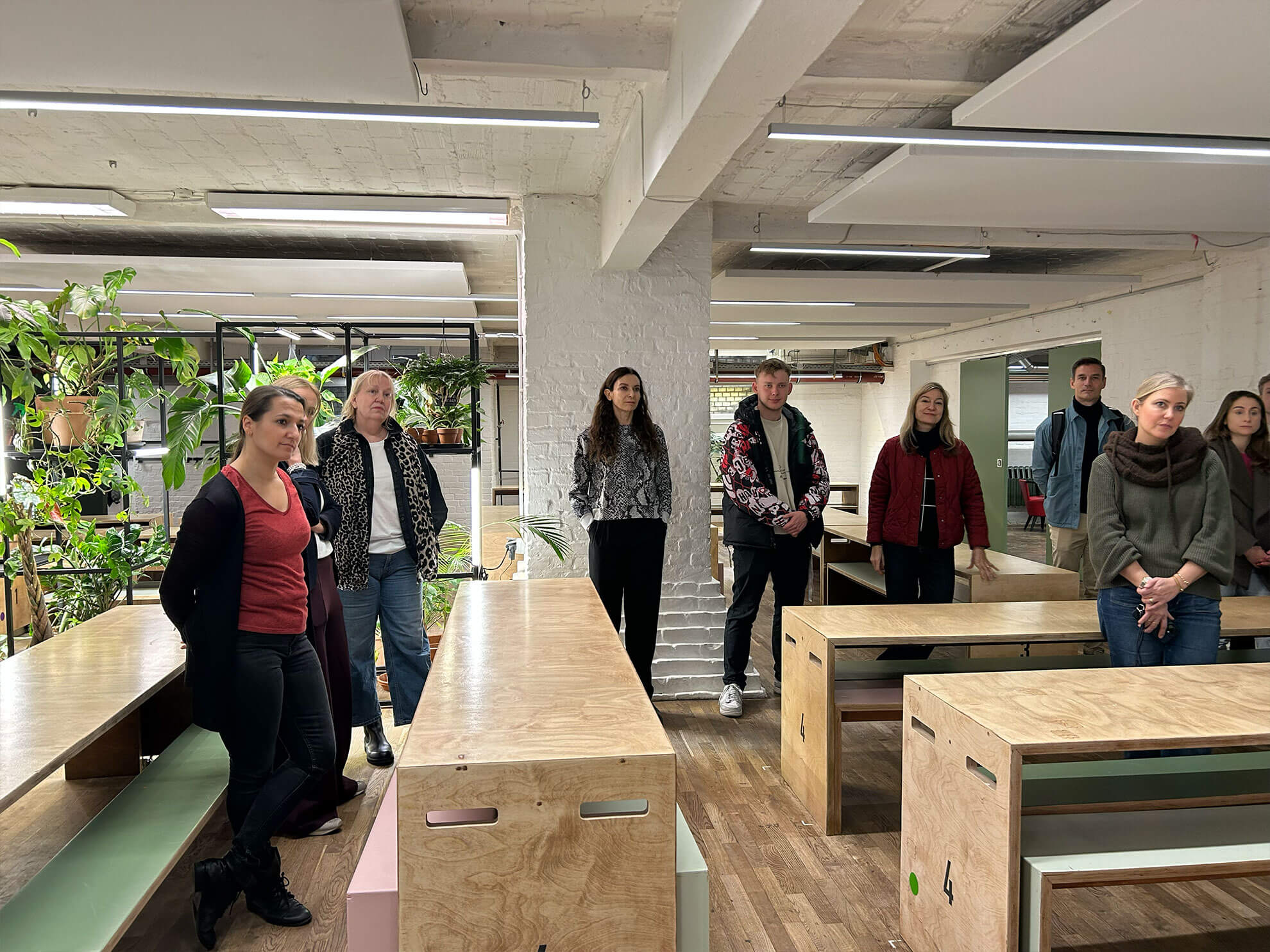 Cosmopolitanschool Berlin Eine Gruppe von Menschen steht in einem modernen Büroraum mit Holzschreibtischen, Pflanzen und weißen Backsteinwänden und hört aufmerksam jemandem zu, der nicht im Bild ist. loading=