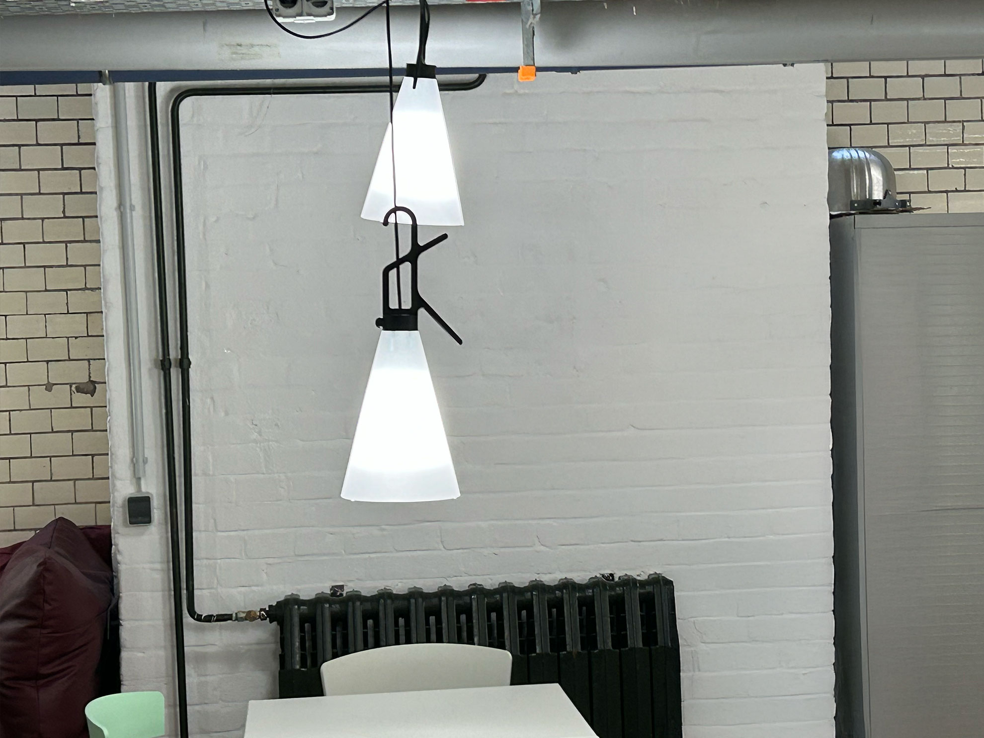 Cosmopolitanschool Berlin Zwei moderne Pendelleuchten mit konischen weißen Lampenschirmen hängen über einem weißen Tisch und Stühlen in einem hellen Raum mit weißen Backsteinwänden und freiliegenden Rohren. An der Wand ist ein schwarzer Heizkörper zu sehen.