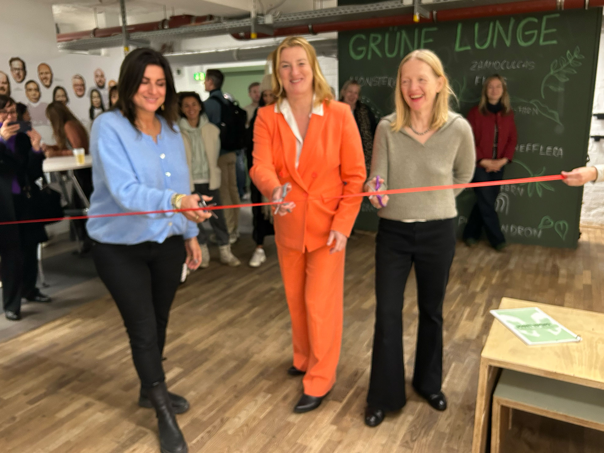 Cosmopolitanschool Berlin Drei Frauen lächeln und durchschneiden ein rotes Band bei einer Indoor-Veranstaltung, mit Schaulustigen hinter ihnen. Im Hintergrund ist ein grünes Schild mit der Aufschrift GRÜNE LUNGE zu sehen, und die Menschen sind versammelt, um den Anlass zu feiern.